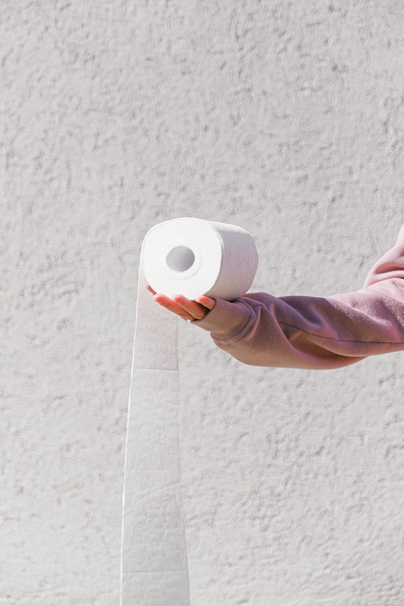 A person in a pink sweater holds a toilet paper roll against a textured wall background.
